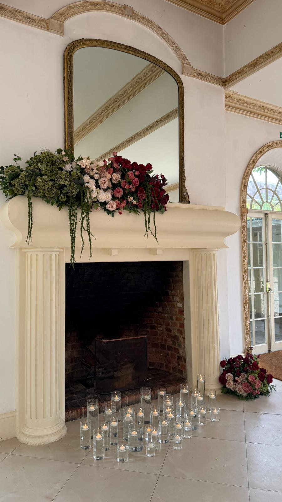 A garland of hydrangea and burgundy roses along a cream mantel, with floating candles on the hearth