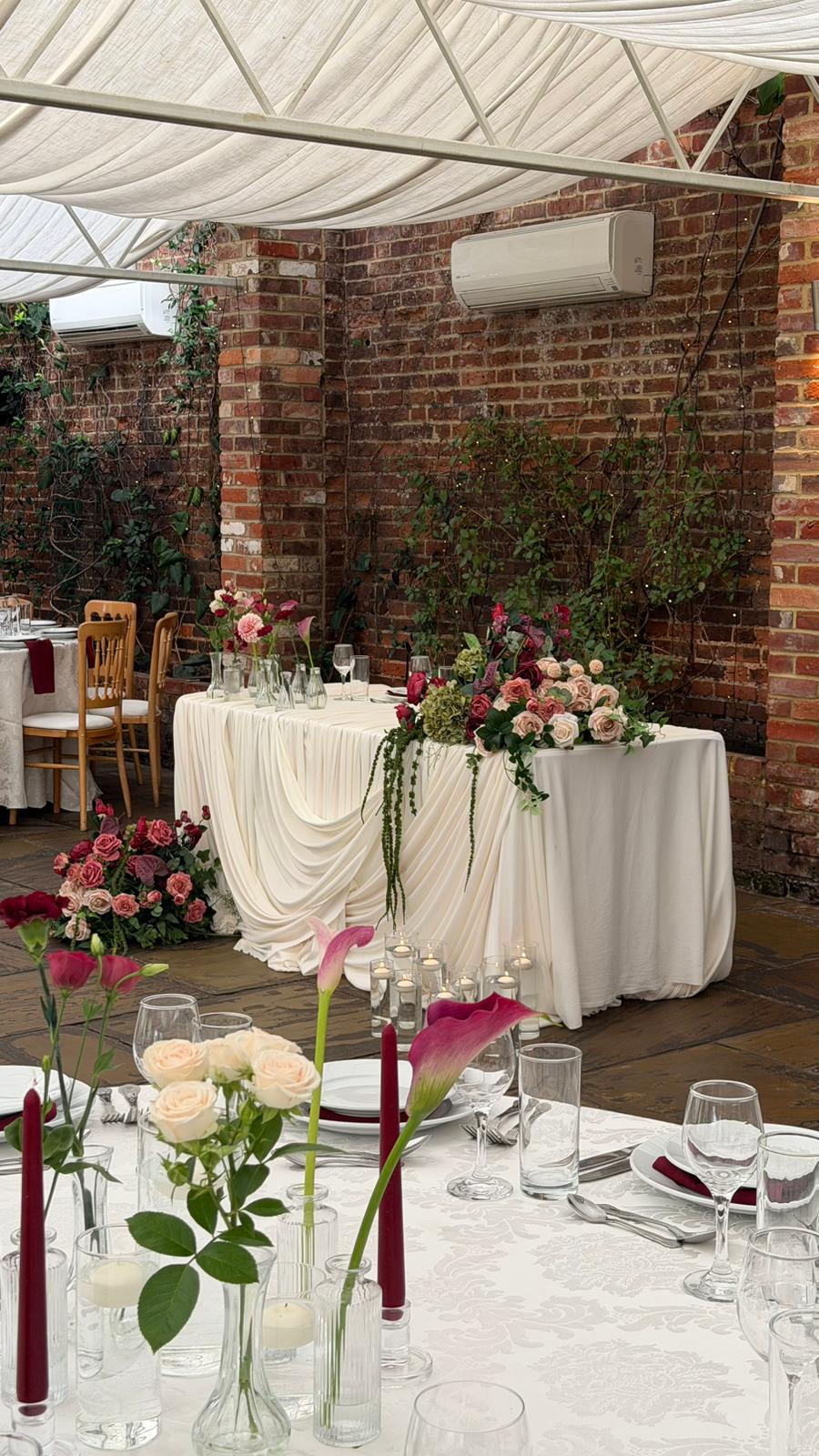 A head table in ivory drape with a swag of blush and burgundy roses in front of a brick wall